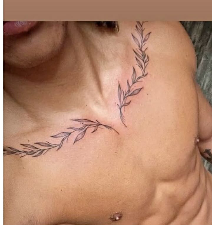 Leafy branch tattoos over both collarbones with a small cross in the center of a man’s chest