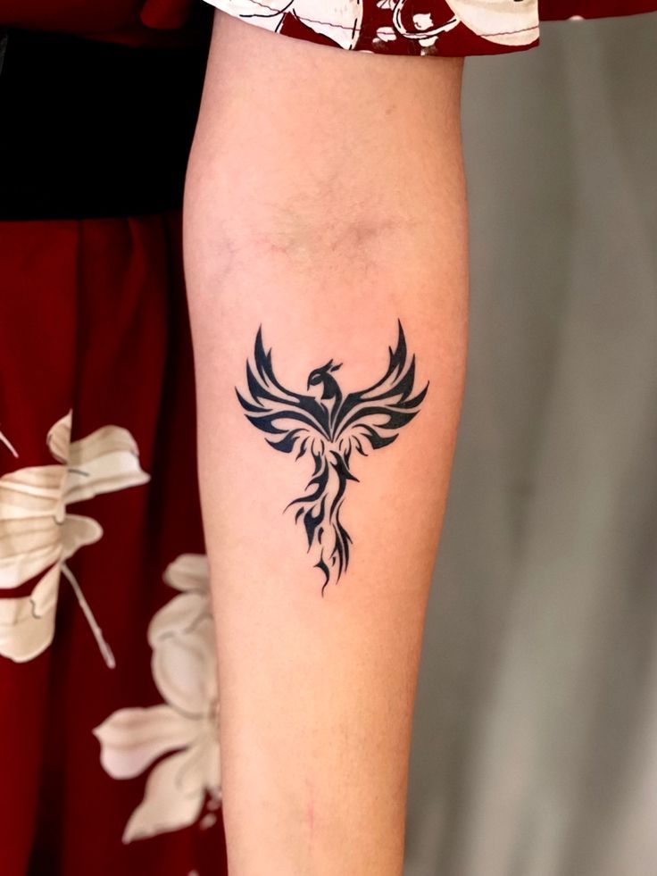 Text: Small black tribal phoenix tattoo with spread wings on a forearm