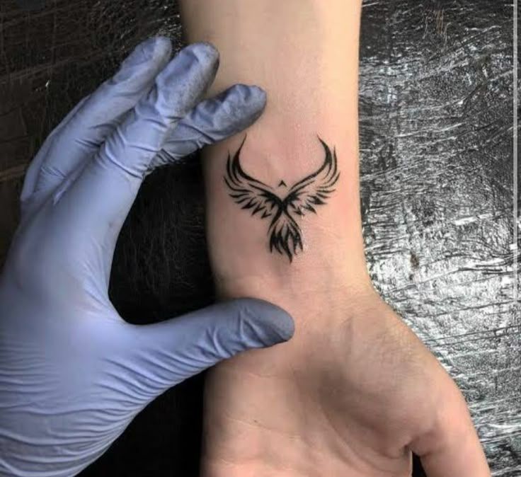 Small black phoenix wings tattoo on the inner wrist while the tattoo artist holds the arm with a glove