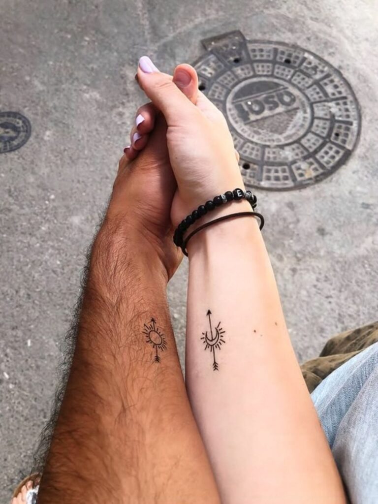 Sun and moon matching tattoos with arrows on a couple’s forearms as they hold hands