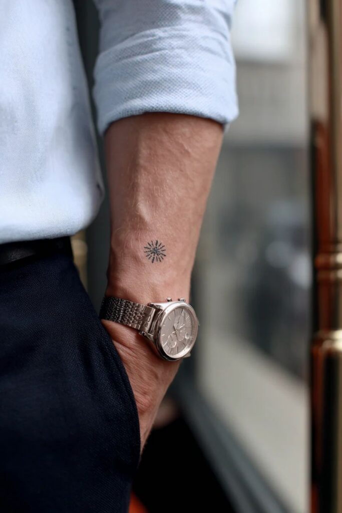 Tiny sun or star tattoo on the forearm of a man wearing a light blue shirt and a silver watch