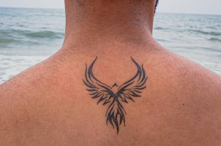 Tribal-style black phoenix with spread wings tattooed at the base of the neck
