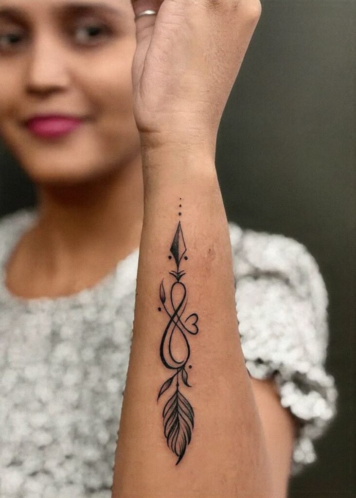 Vertical forearm tattoo of an arrow, infinity loop, small heart and feather