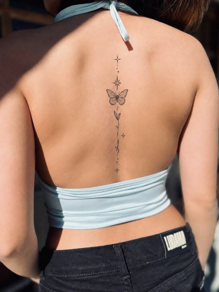 Fine-line butterfly spine tattoo with stars and a leafy stem on a woman’s back in a light blue halter top