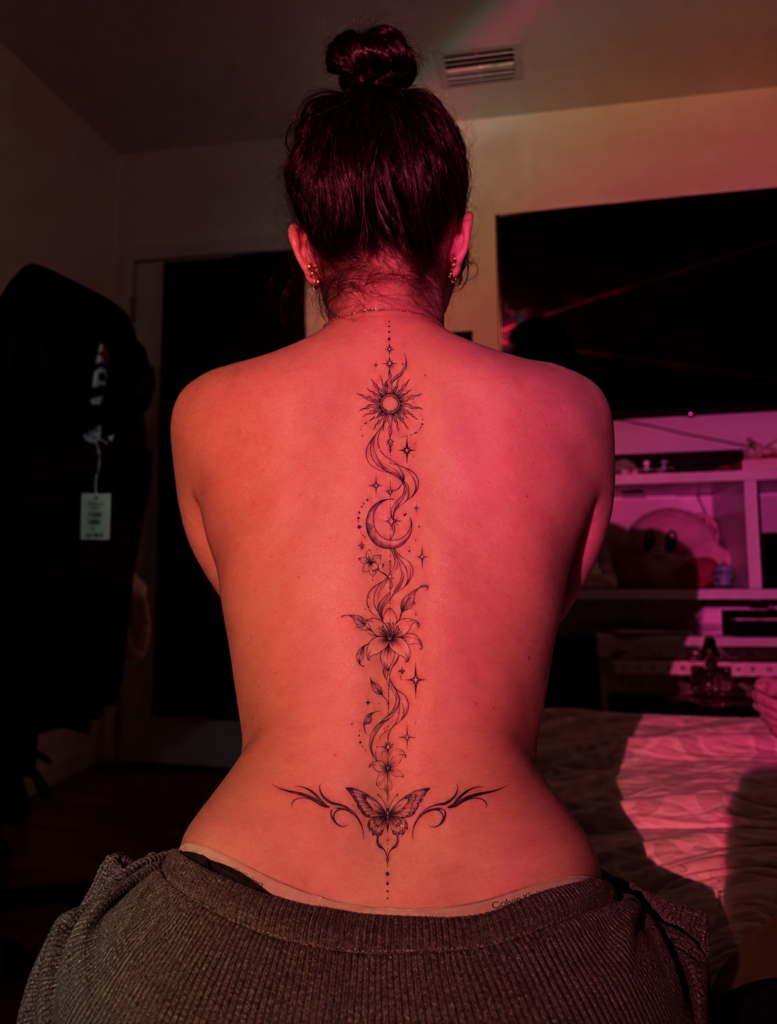 Fine-line sun and moon spine tattoo with lily flowers and butterfly on a woman with her hair in a bun