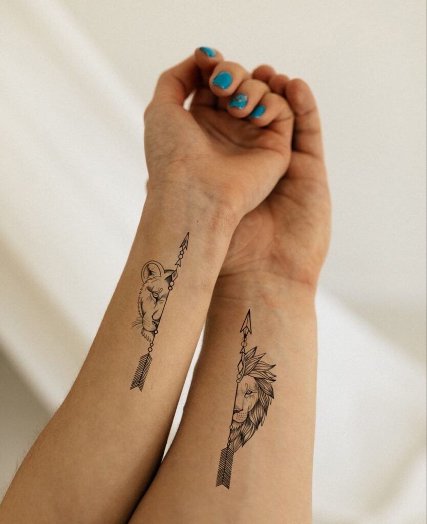 Matching lion and lioness arrow tattoos on two forearms held up together