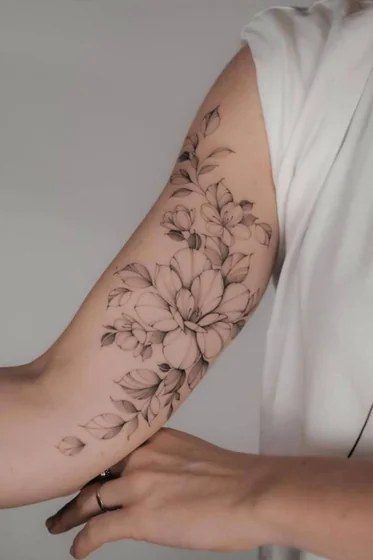 Pink and red painterly flower tattoo with dark green leaves on a woman’s upper arm