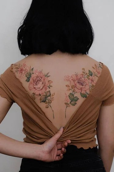Pink rose and wildflower tattoos in color across both shoulder blades on a woman’s upper back
