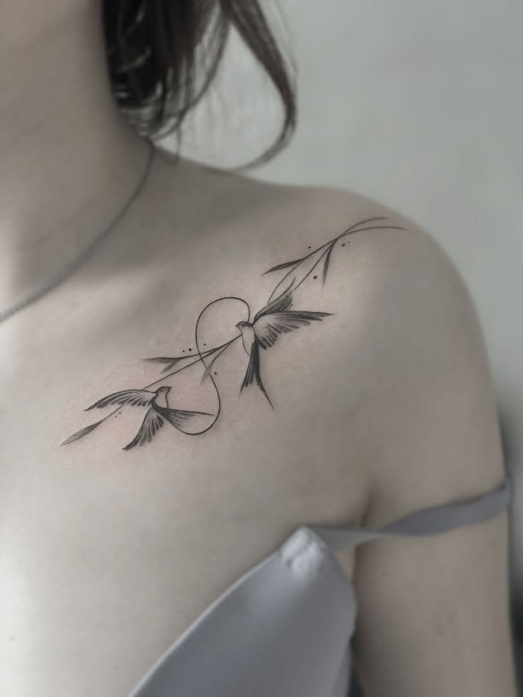 Two swallows connected by an infinity loop and a leafy branch tattoo on the collarbone and shoulder