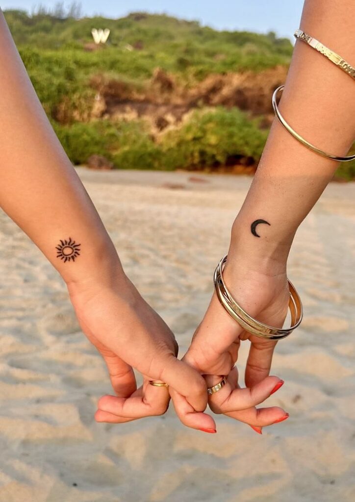 Tiny matching sun and crescent moon wrist tattoos on two people holding hands on a beach